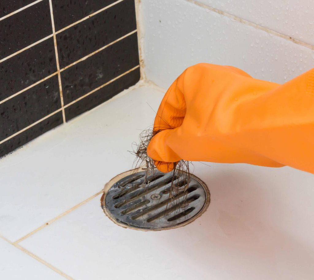 Person wearing an orange rubber glove removing hair clog from a bathroom floor drain during routine cleaning.