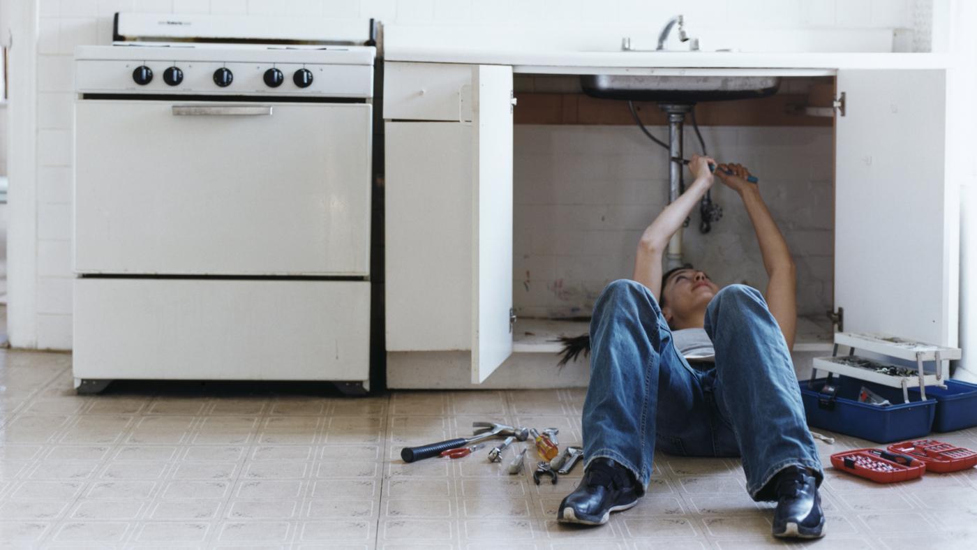 Plumber lying on floor fixing leaking pipes under a kitchen sink in a residential home with tools scattered nearby.